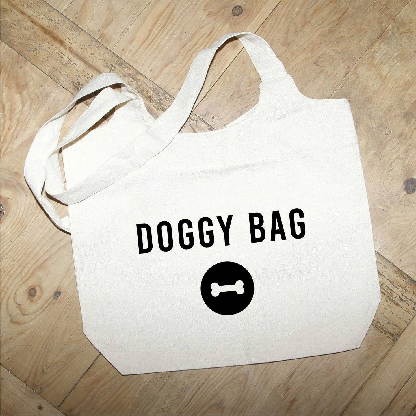 Off white cotton tote bag with DOGGY BAG text and small black bone icon on a wood floor background