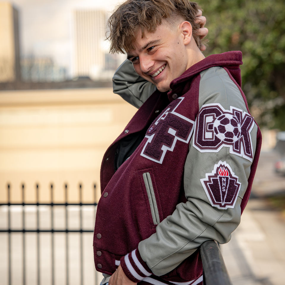 High school senior leaning casually against a fence in urban Houston, proudly wearing his maroon letterman jacket and showing off achievement patches — photographed by Endearing Reflections, a leading high school graduation photographer near The Woodlands, TX.