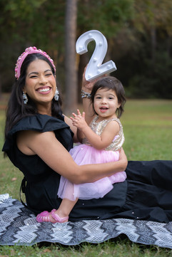 Spring, TX family photography session — mother and daughter sharing a joyful moment on a picnic blanket in a local park. The mom holds a '2' prop above her child, highlighting her age, amidst a natural, leafy backdrop. Captured by Endearing Reflections, renowned for family portraits in Spring, Texas.