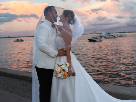 Bride and groom standing together under the bride's veil beside a Florida waterfront at sunset.