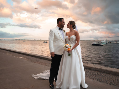 Bride and groom in formal attire stand by the waterfront at sunset, gazing at each other. Sky is colorful; boats are in the background.