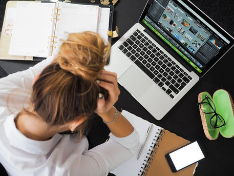 view from a desk where a woman with hair in a bun has her head in her hands. The desk has two open notebooks and an open laptop.