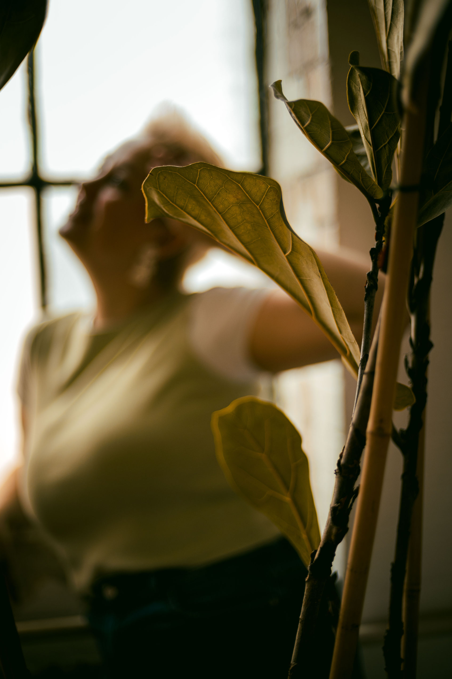 Photo of a woman with short, spiky blond hair. She is out of focus, there are leaves from a potted plant in front of her.