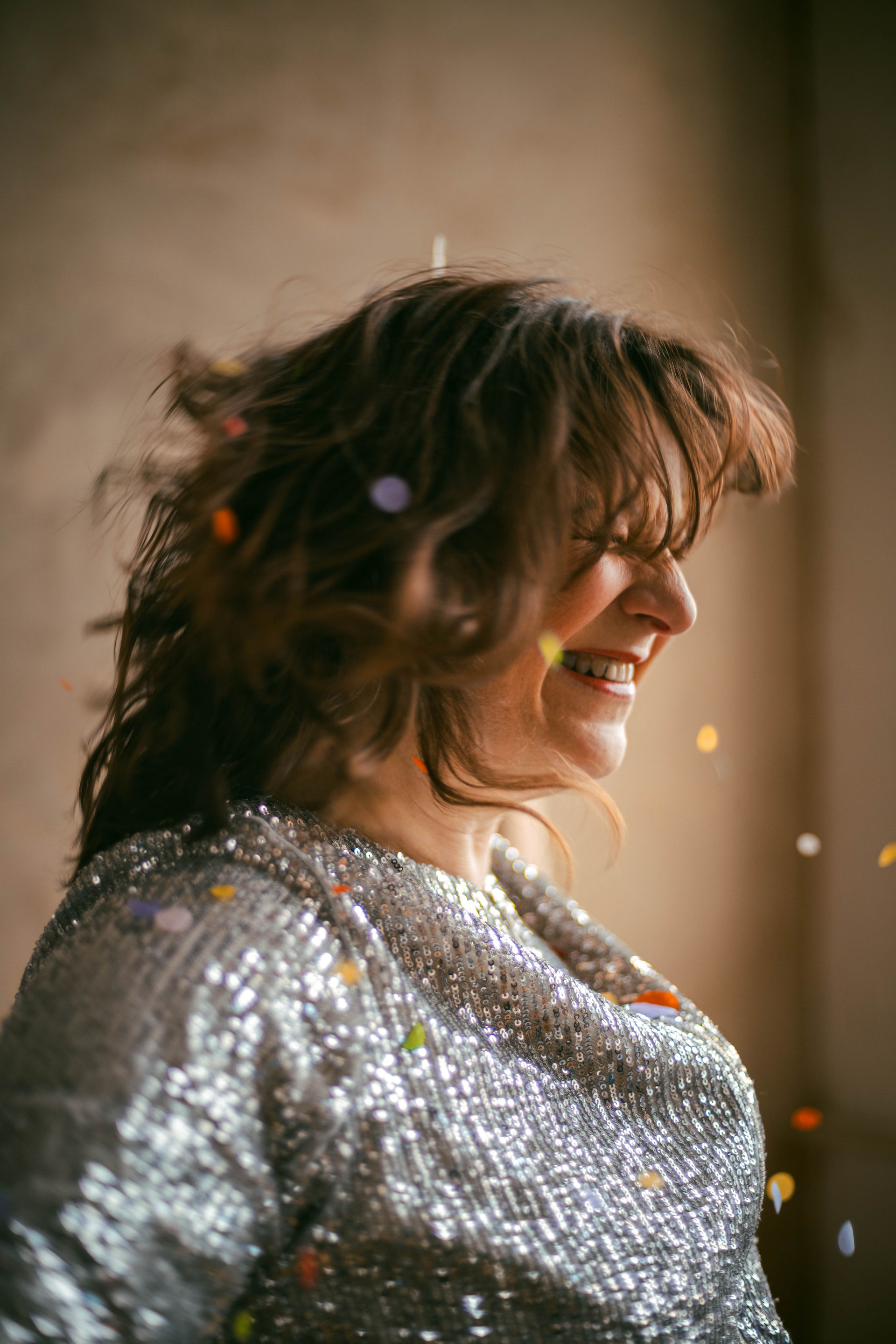 Photograph of a women with shoulder length brown hair, red lipstick, and a silver sequin top. She is spinning, her hair bouncing, and confetti falls out of her hair. The confetti is colourful.