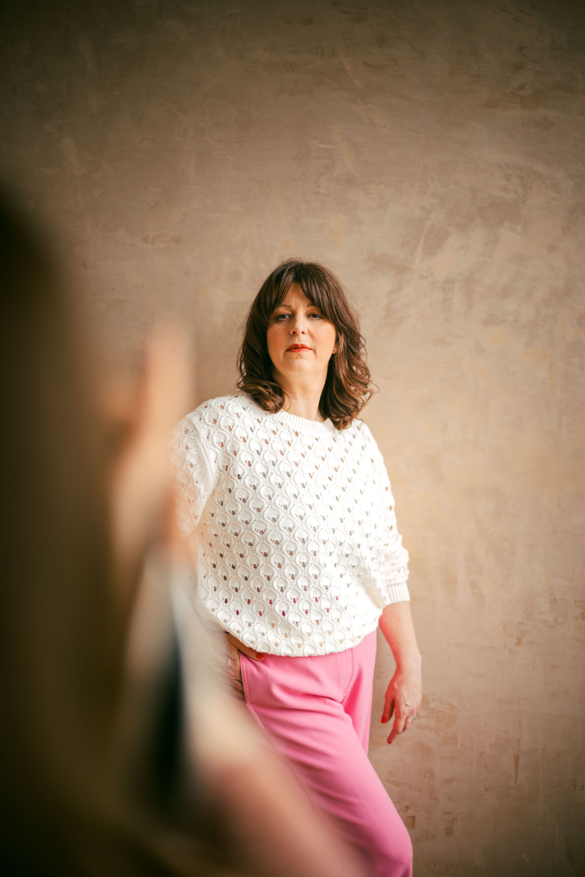 Photograph of a women with shoulder length brown hair, red lipstick, a white knitted jumper and pink trousers. She poses for a photographer in front of a tan, plastered wall.