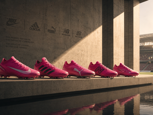 Primer plano de varios tenis de fútbol de color rosa fucsia de distintas marcas alineados sobre el césped de un estadio.