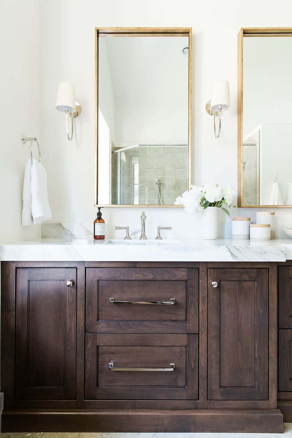 Elegant bathroom with dark wood vanity, marble countertop, two mirrors, gold accents, and white flowers in a vase. Bright and serene.