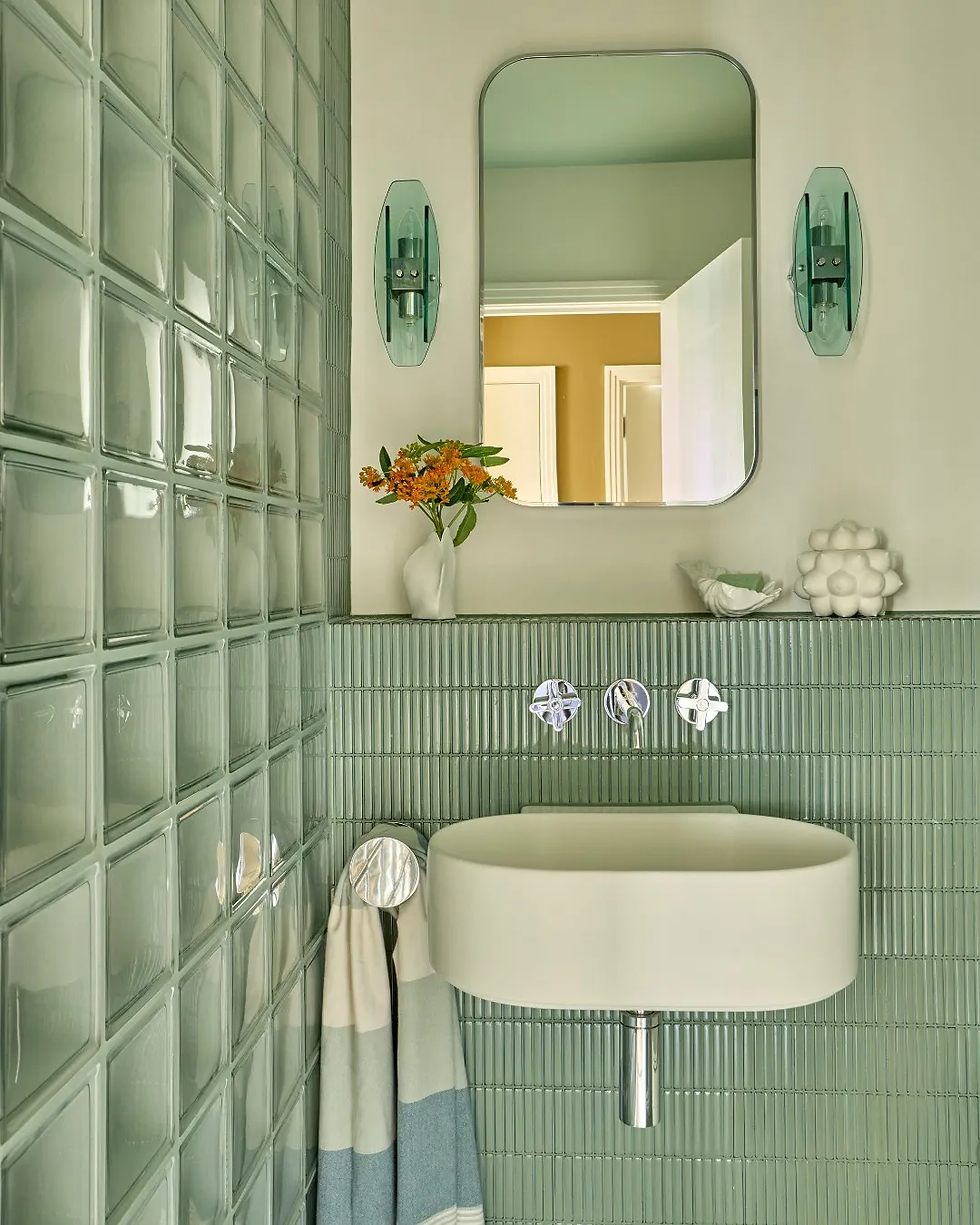 Modern bathroom with green tiles, white sink, and a mirror. Orange flowers in a vase on the shelf. Blue and white towel hangs nearby.