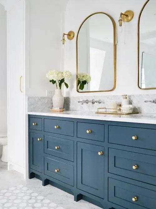 Blue vanity with gold knobs, double mirrors, and sconces in a bathroom. White flowers in a vase, marble countertop, elegant decor.
