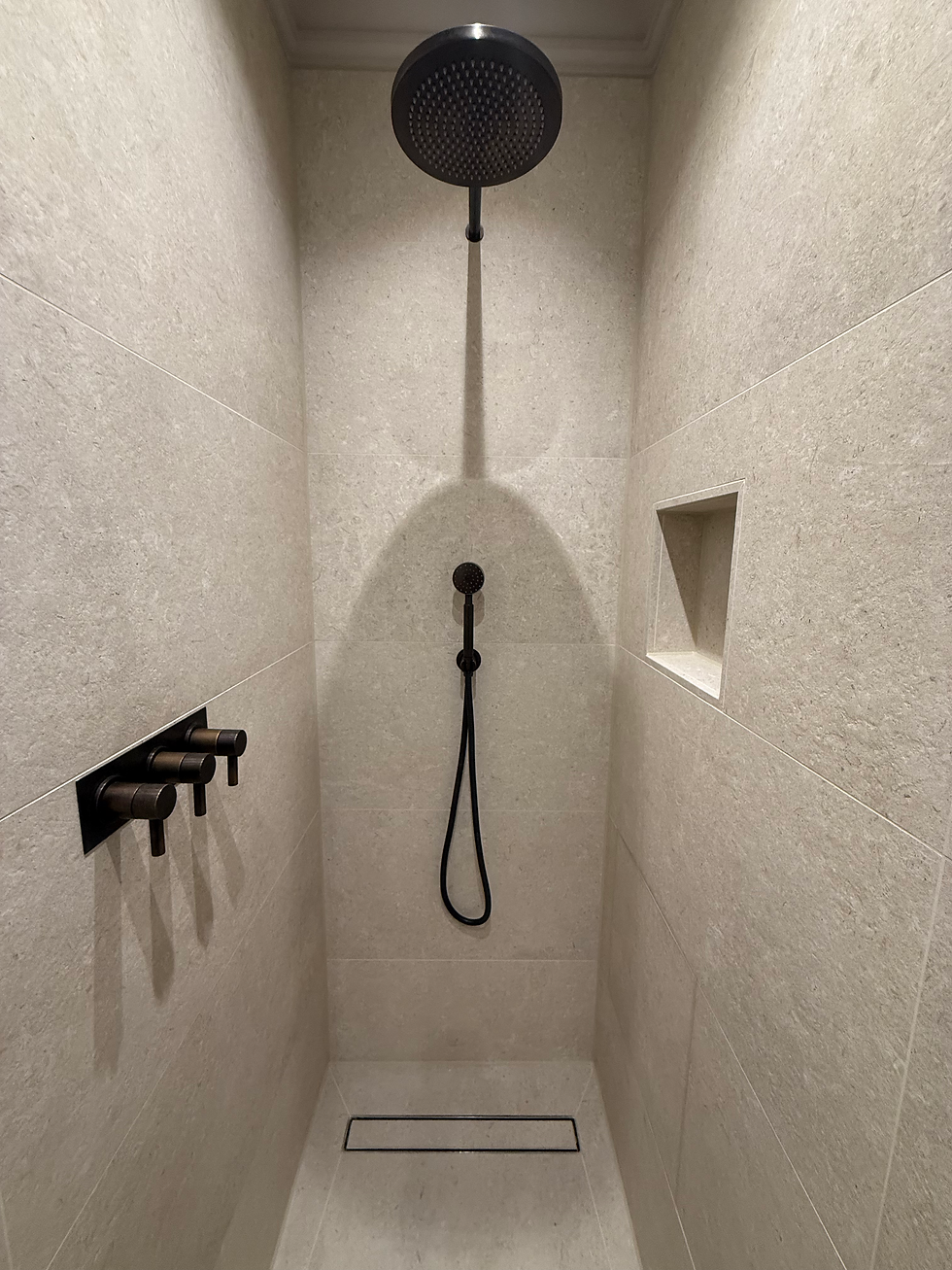 Minimalist beige shower with a black rain showerhead, hose, and controls. A niche on the wall adds storage. Simple and modern design.
