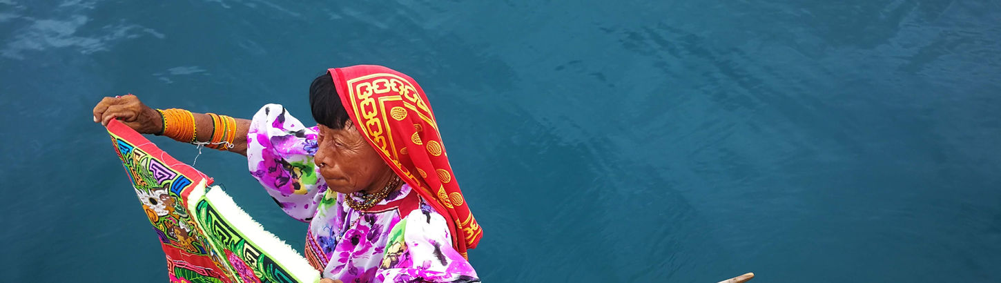 A woman wearing a red headscarf and colorful clothing paddling a small wooden boat on blue water, likely selling goods or navigating a floating market.
