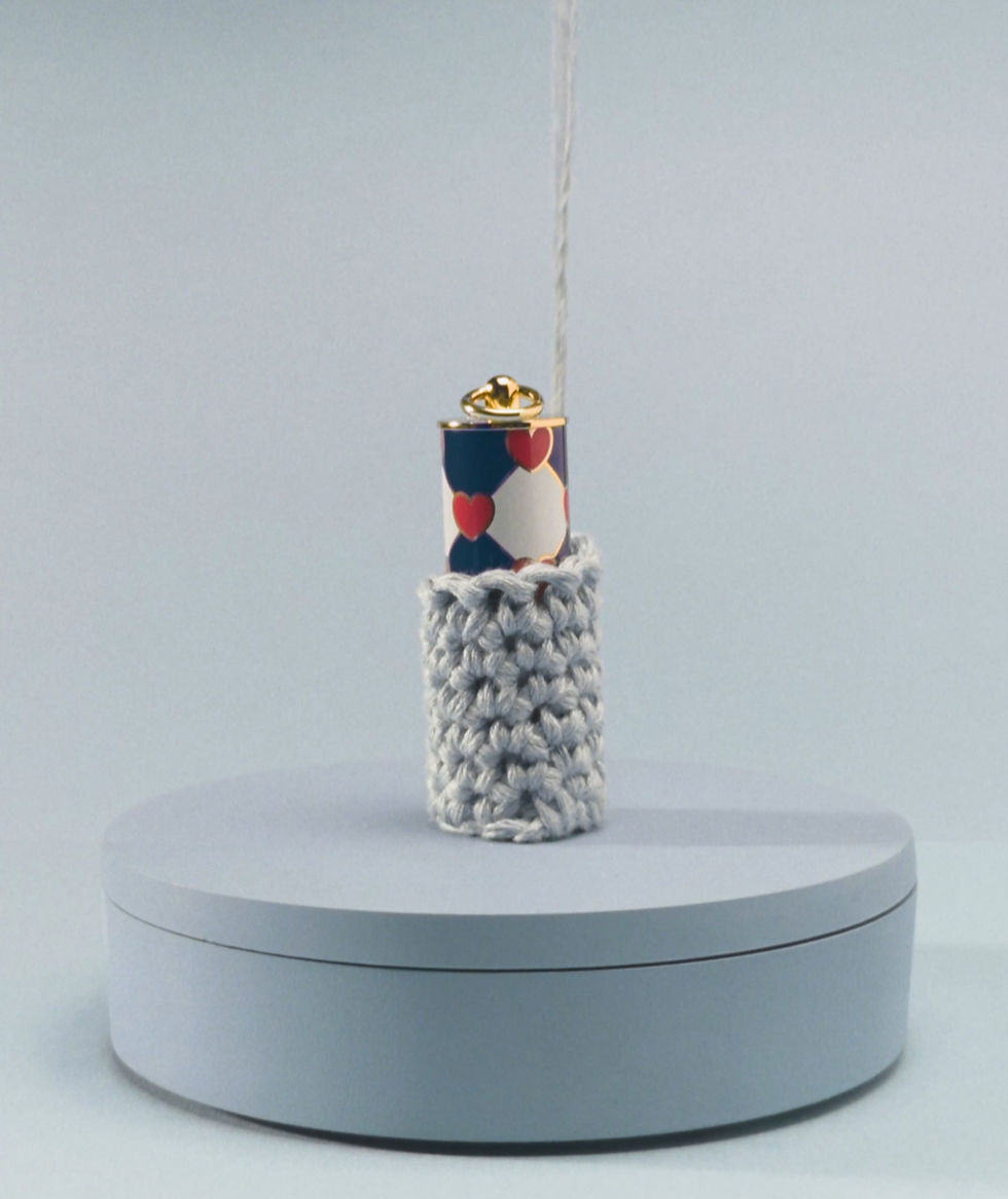 Crochet sleeve slowly pulled upward by a thread to reveal a Carolina Herrera lipstick with heart pattern, on a pale blue pedestal.