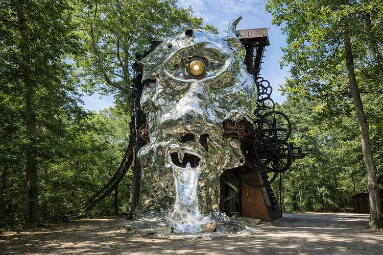 Niché au cœur du "Bois des Pauvres", à la sortie de la ville, se cache l'une des œuvres d'art contemporain les plus monumentales et insolites d'Europe : le Cyclop.
Une Œuvre Monumentale et Collective : Conçue en secret à partir de 1969 par le sculpteur suisse Jean Tinguely et sa compagne Niki de Saint Phalle, cette sculpture-architecture mesure plus de 22 mètres de haut et pèse environ 350 tonnes d'acier et de béton. Elle a réuni les contributions d'une quinzaine d'artistes amis (comme Daniel Spoerri ou César).
Une Structure Rutilante : Le monstre possède un œil unique doré, une oreille articulée d'une tonne et une immense face recouverte de milliers de miroirs qui reflètent la forêt environnante, intégrant parfaitement l'art sauvage à la nature. Sa langue est une immense glissière (toboggan) métallique.
Un Labyrinthe Mécanique Intérieur : Lors des visites guidées (accessibles dès 8 ans), on pénètre à l'intérieur de la tête pour découvrir un univers surréaliste. Les engrenages de ferraille s'animent dans un fracas métallique, des billes géantes circulent sur des rails, et de nombreuses œuvres d'art clandestines (dont un hommage à Yves Klein au sommet) ponctuent les différents étages.