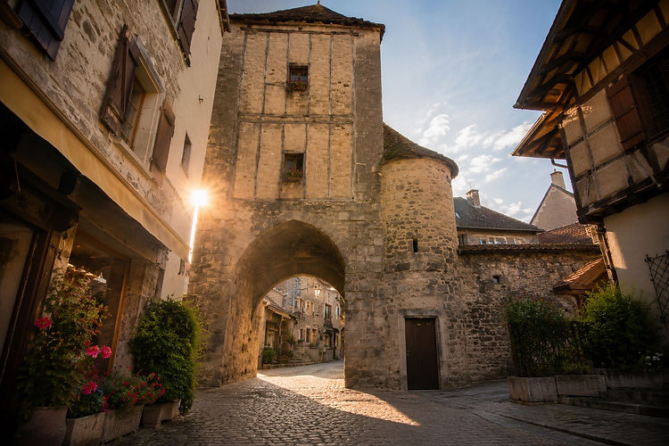 L'exploration de Rocamadour commence au pied de la falaise, là où s'étire l'unique rue médiévale pavée de la cité, autrefois foulée par les rois et les mendiants.
La Porte du Figuier et la Porte Salmon : Ces imposantes portes fortifiées en pierre calcaire marquent l'entrée de la cité. Elles rappellent que Rocamadour, outre sa vocation religieuse, était une place forte militaire conçue pour résister aux pillards.
La rue de la Couronnerie : Bordée de magnifiques demeures médiévales aux façades de pierre, cette ruelle vivante abrite aujourd'hui des échoppes d'artisans, des terrasses accueillantes et des boutiques de produits régionaux, offrant des points de vue vertigineux dès que l'on lève les yeux vers le sommet.
La Maison de la Pomme de Pin : L'une des plus vieilles demeures du village, remarquable par son architecture typique du Quercy, témoin du quotidien des marchands qui vivaient du commerce généré par l'afflux des pèlerins.