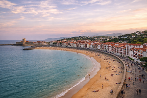 Contrairement à ses voisines de Biarritz ou d'Anglet, la plage de Saint-Jean-de-Luz offre des eaux calmes et sécurisées, grâce à un exploit d'ingénierie maritime unique au monde.

    Les Trois Digues de Socoa, Sainte-Barbe et l'Artha : Au XIXe siècle, la ville fut menacée de destruction complète par les tempêtes hivernales de l’Atlantique. Napoléon III ordonna la construction de trois digues de granit colossales fermant la baie. Ce rempart brise la force des vagues du large, transformant le littoral en un bassin paisible.

    La Grande Plage et les Passerelles Napoléon III : S'étirant sur plus d'un kilomètre de sable fin, cette plage familiale est longée par une magnifique promenade piétonne. Elle est bordée de maisons néo-basques reliées à la digue par d'élégantes passerelles en bois, offrant un cachet architectural "Belle Époque" irrésistible.

    La Pointe de Sainte-Barbe : Situé à l'extrémité nord de la baie, ce promontoire verdoyant est le poumon vert de la ville. Une promenade sur ses sentiers escarpés mène à une petite chapelle de marins et dévoile un panorama époustouflant à 360 degrés sur toute la baie, les vagues de la structure de surf de Belharra au large, et les falaises de la Corniche Basque.