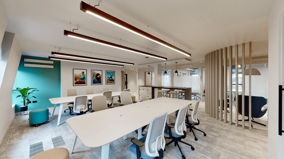 A striking open-plan office desking space with light-coloured desk chairs and black track lights overhead, positioned in front of a wall displaying three colourful photographs.