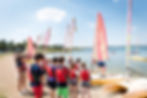 Students in red life jackets gathered by brightly coloured sailing boats on the shore, preparing for a water activity.
