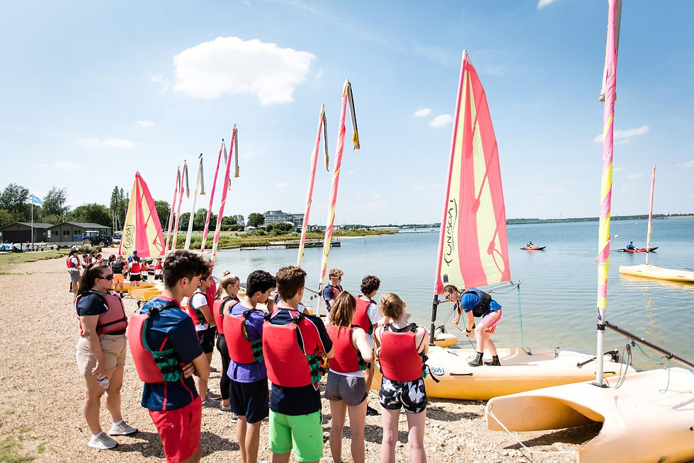 Students in red life jackets gathered by brightly coloured sailing boats on the shore, preparing for a water activity.