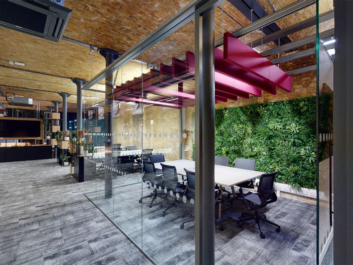 A unique and striking glass-walled meeting room with an open top, suspended linear acoustic panels in a bright reddish-pink hue, and a feature green wall composed of plants.
