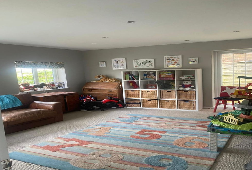 Bright kids’ playroom with brown sofa, toy shelves, stuffed animals, ride-on cars, and colorful number rug by the window