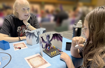 2 players at a game table. Each has a game board in front of them, shielded by dragon themed screens. The brightly colored game pieces contrast against the black game board.