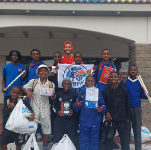 Tony steht lächelnd zwischen einer Gruppe namibischer Schulkinder, die bunte neue Schulrucksäcke tragen und Daumen nach oben zeigen.