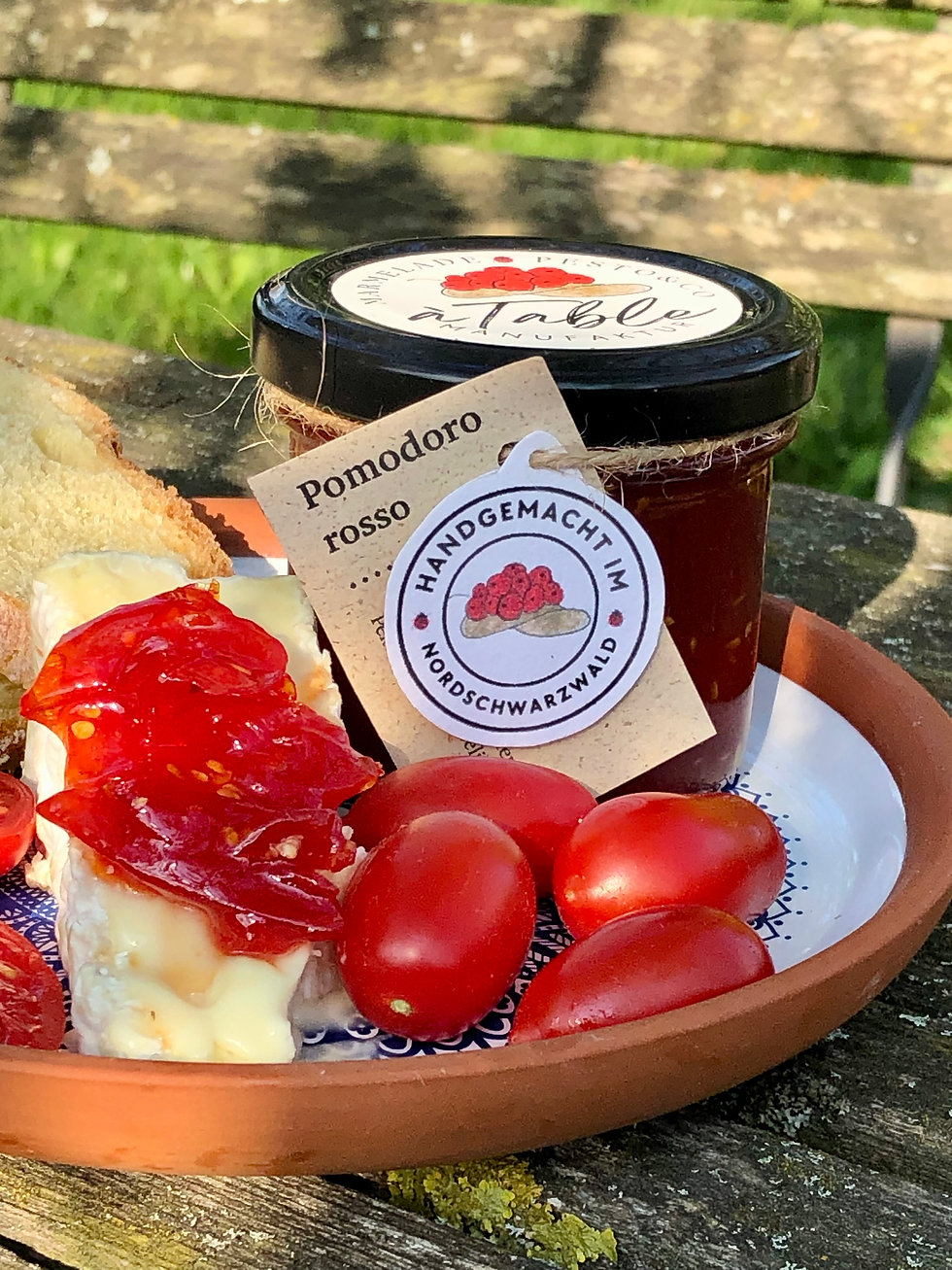 Ein Glas Tomaten-Fruchtaufstrich Badische Pomodori mit kleinen Tomaten. Etwas Fruchtaufstrich liegt auf Camembert
