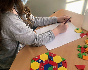 A young female student draws something on a piece of paper with a colorful pattern block design next to her on the table.