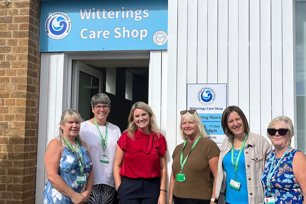 Jess Brown-Fuller and local representatives pose outside Witterings Care Shop under a blue sign, smiling in bright summer clothes and lanyards.