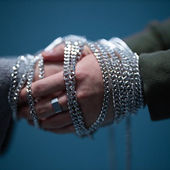 Hands clasped and wrapped in silver chains against a blue background, conveying restraint. One hand wears a ring. Mood is tense.