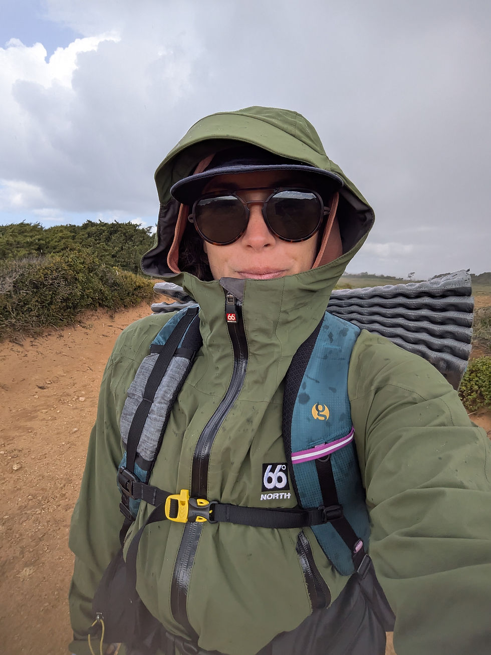 Hiker wearing a rain jacket on the Fisherman’s Trail, navigating changing weather with cloudy skies and coastal terrain in Portugal.