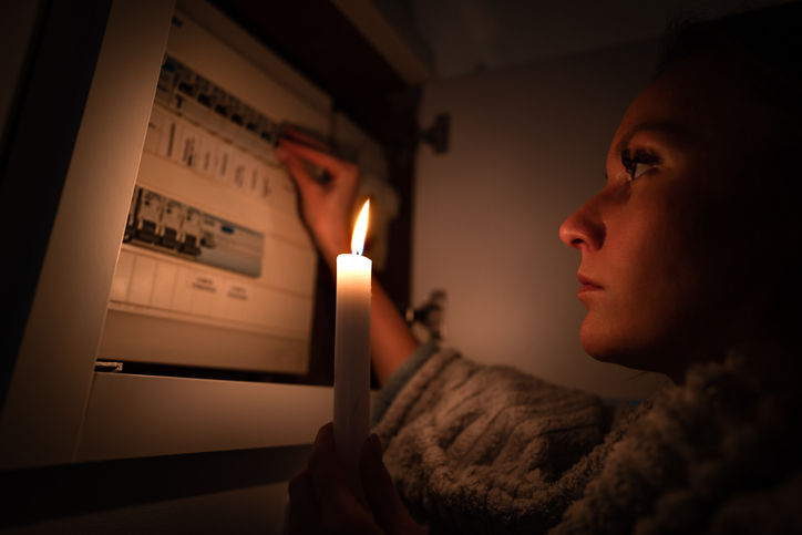 Woman holding a lit candle while inspecting a circuit breaker during a power outage, highlighting the vulnerability of unreliable backup systems.
