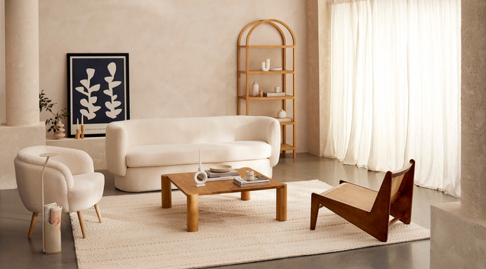 A spacious living room filled with soft, textured furniture in neutral tones, including a bouclé fabric sofa and armchair, a wooden coffee table, and a woven rug, all bathed in natural light from large windows, creating an airy and inviting atmosphere.