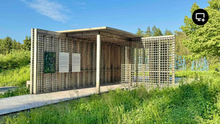 Structure en bois ajourée au design géométrique, installée en pleine nature sous un ciel dégagé. Trois panneaux d’exposition sont fixés à la paroi, dont un affiche le mot « Frontières ». À l’arrière-plan, des structures sculpturales bleues émergent de la végétation haute.