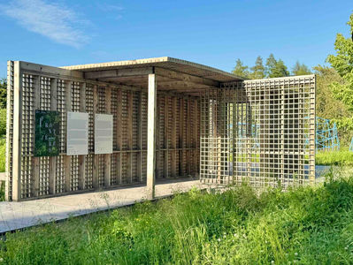 Structure en bois ajourée au design géométrique, installée en pleine nature sous un ciel dégagé. Trois panneaux d’exposition sont fixés à la paroi, dont un affiche le mot « Frontières ». À l’arrière-plan, des structures sculpturales bleues émergent de la végétation haute.