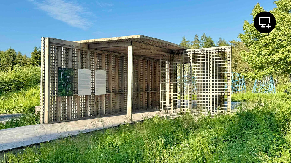 Structure en bois ajourée au design géométrique, installée en pleine nature sous un ciel dégagé. Trois panneaux d’exposition sont fixés à la paroi, dont un affiche le mot « Frontières ». À l’arrière-plan, des structures sculpturales bleues émergent de la végétation haute.