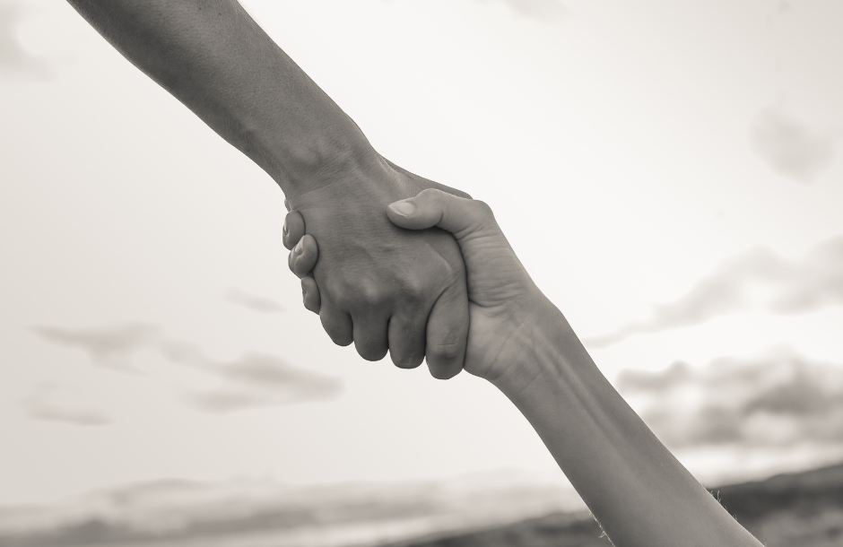 Two hands grasp each other firmly against a cloudy sky backdrop, evoking a sense of support and unity in a sepia-toned image.