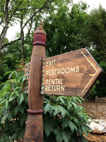 Wooden arrow-shaped wayfinding sign listing exit, restrooms, rental, and return, mounted on a carved post.