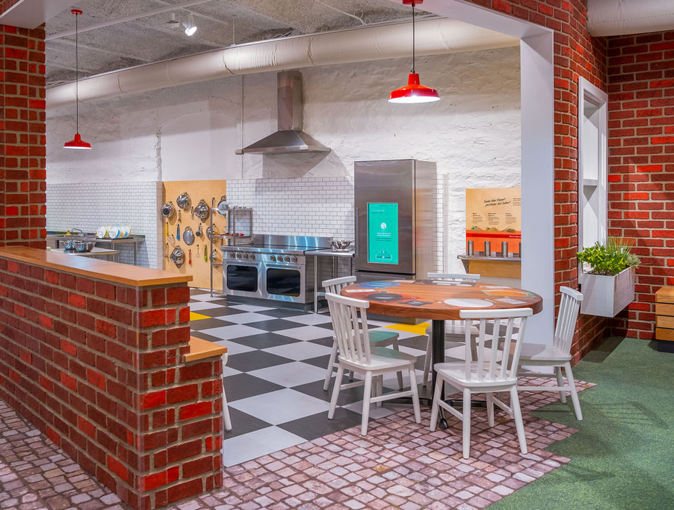 Cozy pretend kitchen area featuring red brick accents, a round dining table, refrigerator display, and hanging lights.