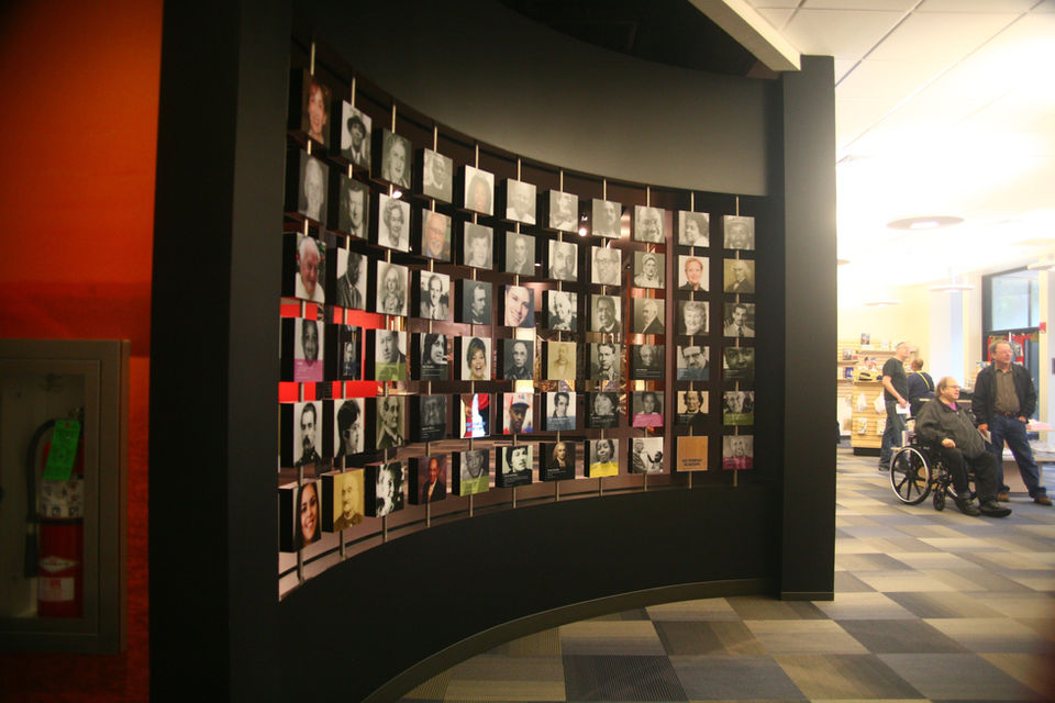 Curved memorial wall filled with portrait photographs of individuals, possibly veterans or community members, within a museum or exhibit space.