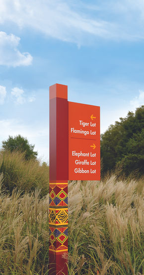 Vertical directional sign listing animal exhibits including tiger, elephant, and gibbon, in front of tall grasses.