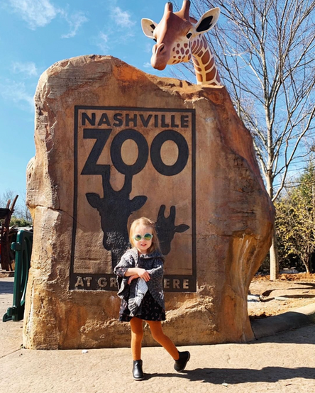 Young child standing in front of a large rock entrance sign reading “Nashville Zoo at Grassmere” with a giraffe sculpture.