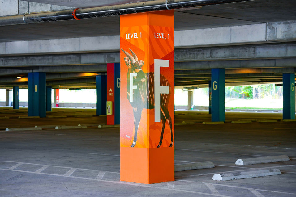 An orange column in a zoo parking garage is labeled “LEVEL 1” with large white letters “F” on multiple sides. The column features a graphic of a bongo antelope, and additional color-coded columns marked with other letters, like “G,” are visible in the background. The structure is well-lit and organized with clearly marked parking areas.