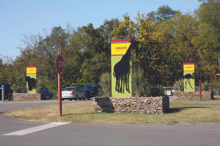 Wide view of the zoo entrance drive with stop signs and animal habitat signs mounted on stone bases.