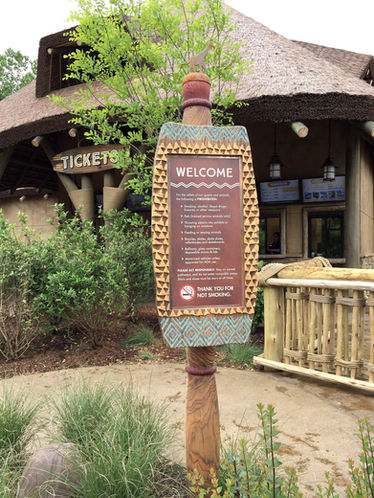 Decorative vertical welcome sign outside a ticketing area, mounted on a carved wooden post.