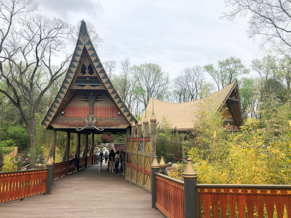 Visitors walking across a red wooden bridge toward Thai-inspired buildings surrounded by trees.