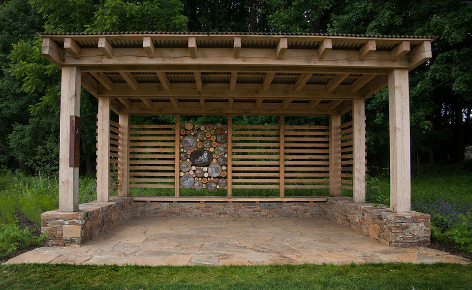 Open wooden shelter with stone base and pollinator wall – A shaded pavilion structure with wood slat walls and a central insect habitat wall stands at the edge of a forest.