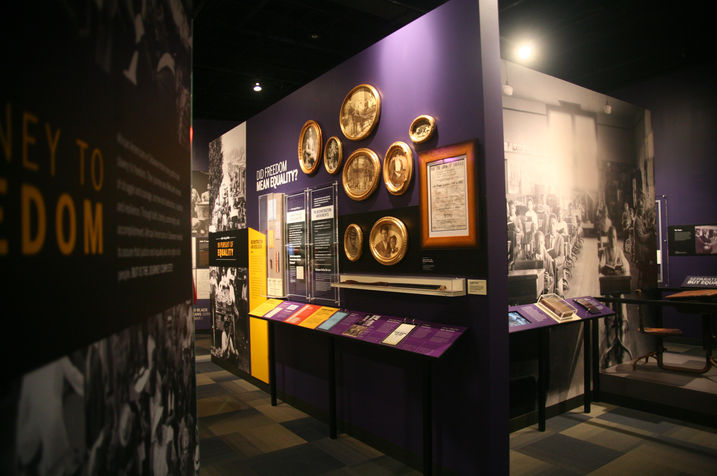 "Journey to Freedom" exhibit panel in front of a purple wall filled with historic photographs and gold-framed portraits highlighting African American history.