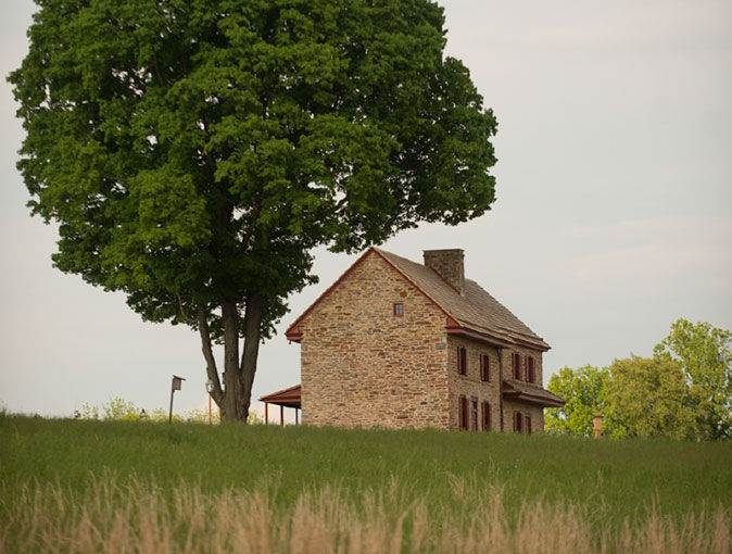 Stone farmhouse beside a tree – A historic stone farmhouse with a large shade tree beside it sits peacefully in a grassy field under a clear sky.