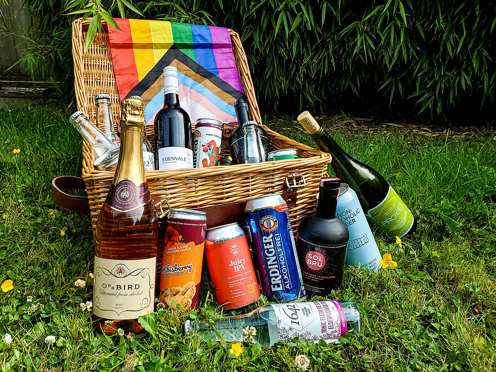 a picnic basket sitting on a lawn with buttercups and clovers,filled with bottles of alcohol-free beverages including beer, wine, and soft drinks. A pride flag drapes over the basket lid.